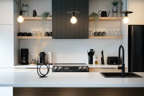 Modern kitchen with white countertop, dark stovetop, open shelving with plants, pendant lights, and organized appliances and dishware