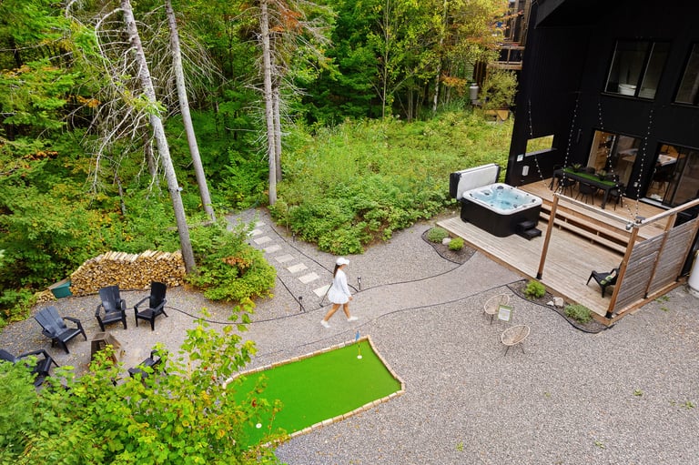 Aerial view of a modern backyard with a bright green lawn, dark house, wooden deck with hot tub, seating area, and surrounding forest trees