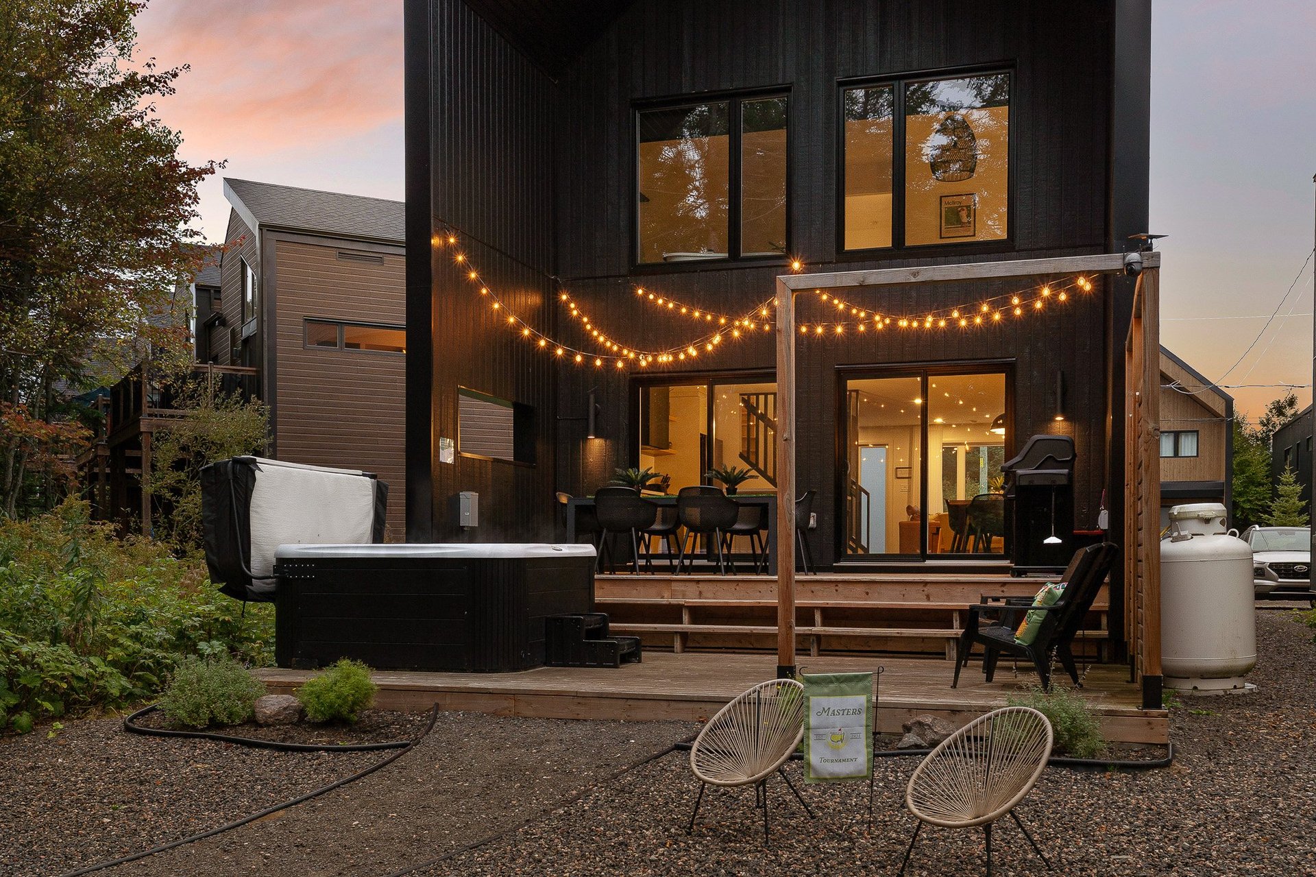Modern black tiny house at dusk with warm string lights, deck seating, and bicycle in front yard at sunset