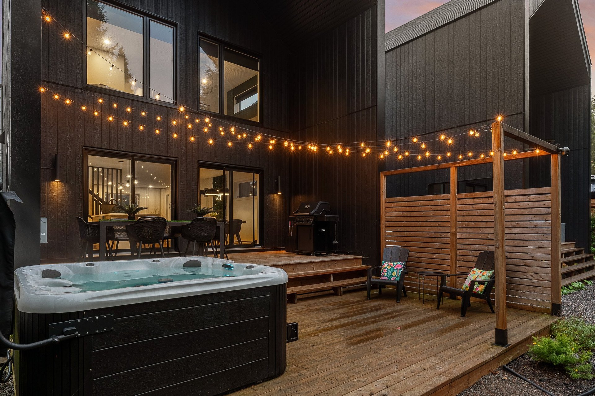 Modern backyard patio at night with hot tub, wooden deck, lounge chairs, pergola, and string lights against dark siding and glass doors
