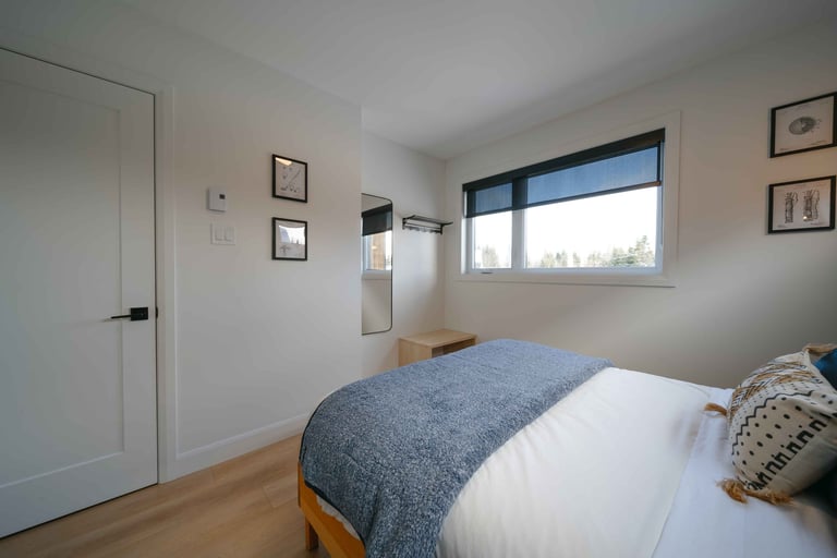 Modern bedroom with wooden bed, blue-gray bedding, white walls, wooden flooring, white door, large window with blue frame, and wall-mounted artwork