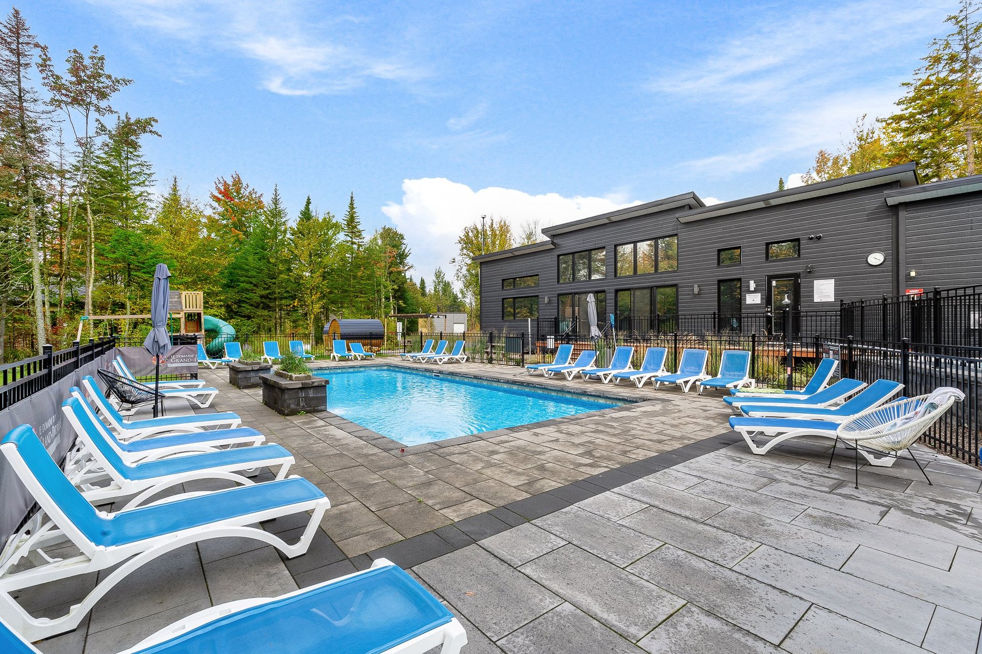 Outdoor resort pool surrounded by blue lounge chairs, modern black building, forested background, and clear sky