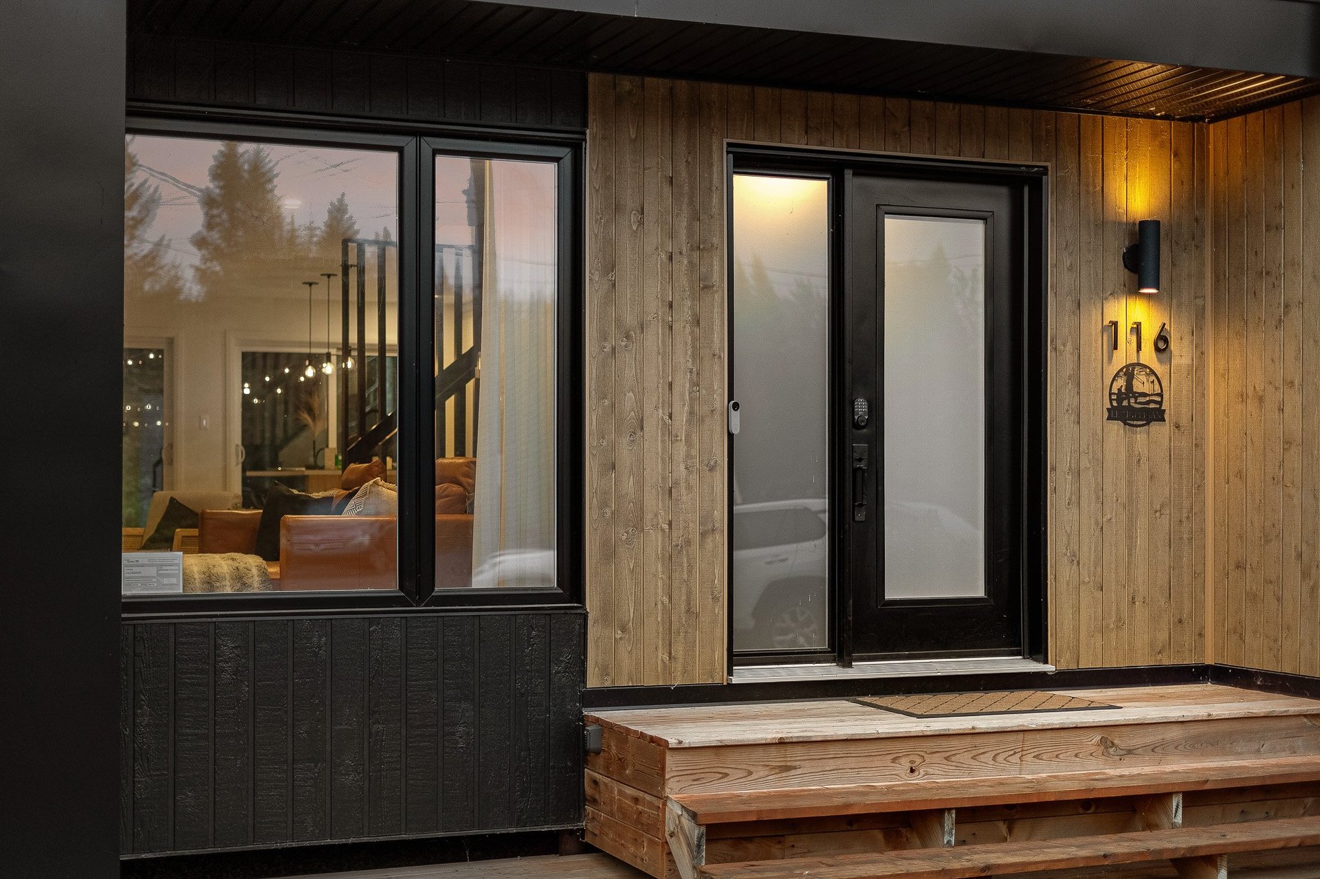 Modern home exterior with wood paneling, black-framed glass doors and windows, warm lighting, and wooden porch deck