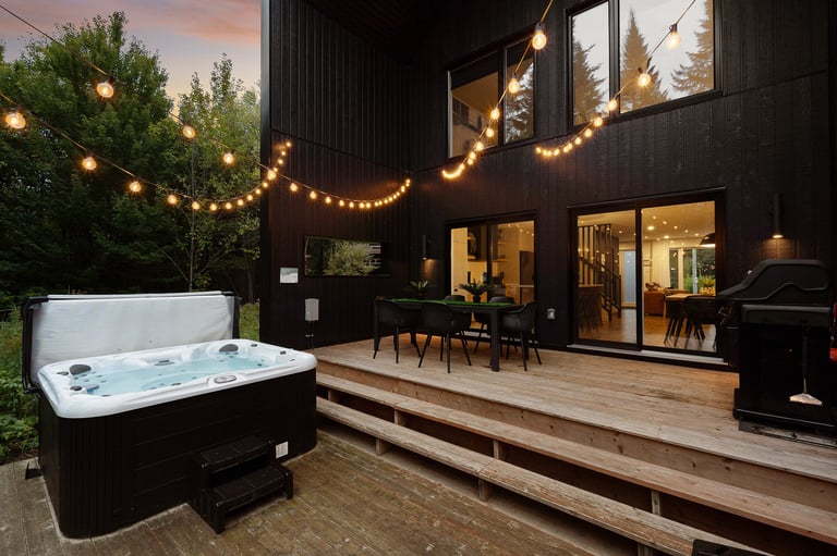 Outdoor deck at dusk with hot tub in foreground, modern black house with illuminated windows, wooden deck, and string lights overhead