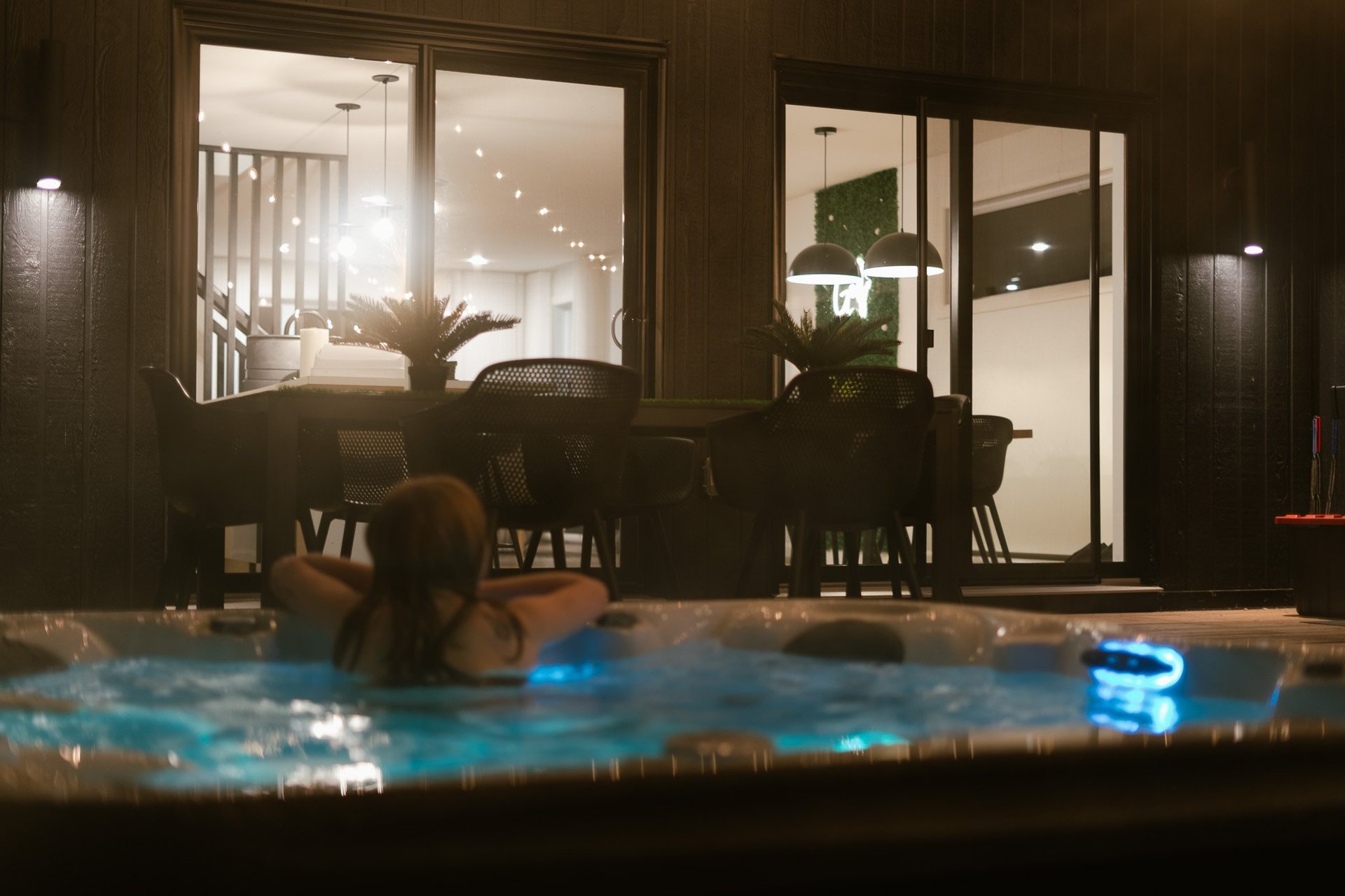 Person relaxing in illuminated outdoor hot tub at night with modern home interior visible through large windows