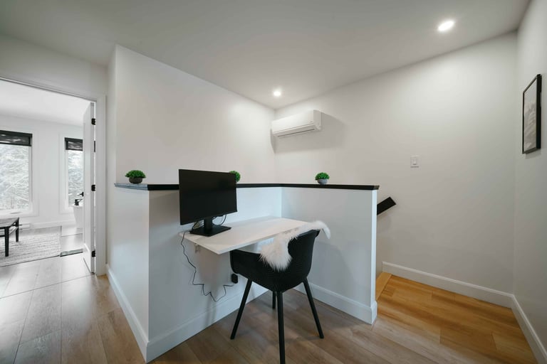 Modern minimalist office space with white walls, built-in desk, black chair, computer monitor, and wooden flooring